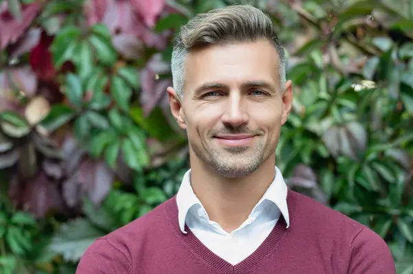 Smiling middle-aged businessman wearing a burgundy jumper and white shirt, standing outdoors with green foliage in the background.